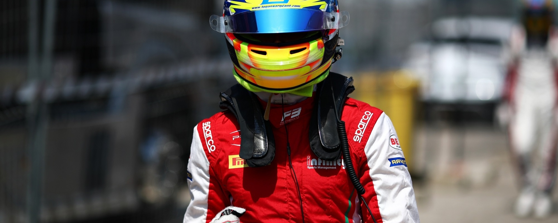 A racing driver in a red and white race suit and a yellow and blue racing helmet pumps his right fist