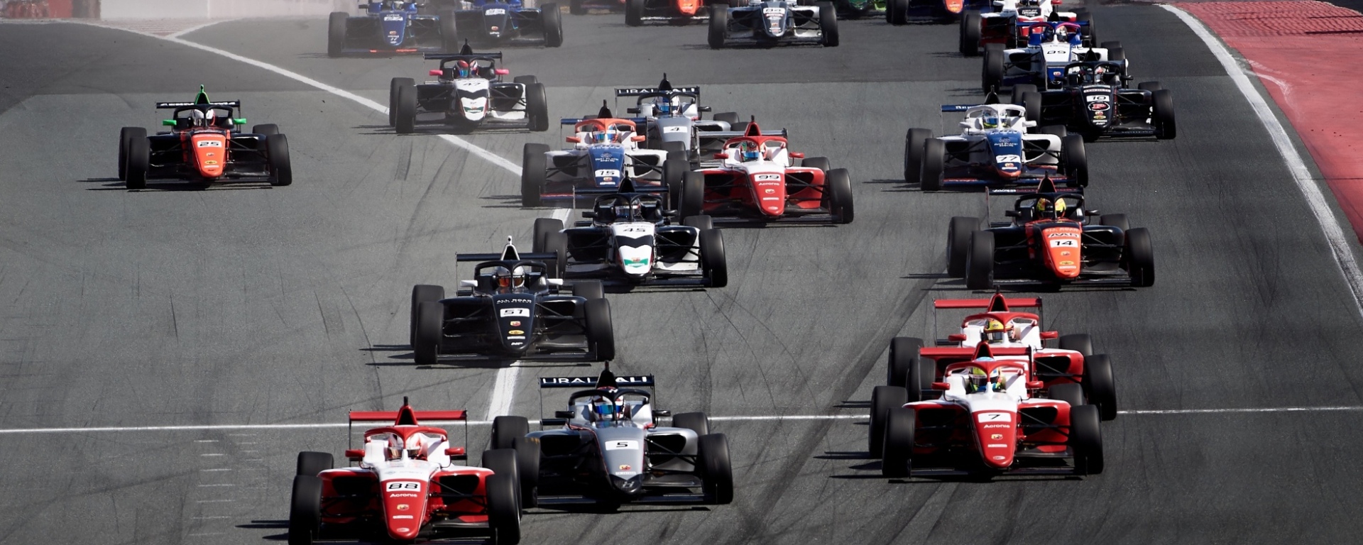 A group of 25+ open-wheel cars heads downhill on a straight