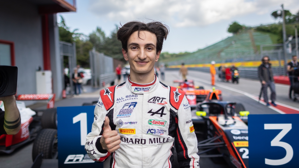 A young man with black hair (Gabriele Minì) in a white, red and black racing suit looks at the camera and does a thumbs-up gesture with his right hand. Several race cars are parked behind him.