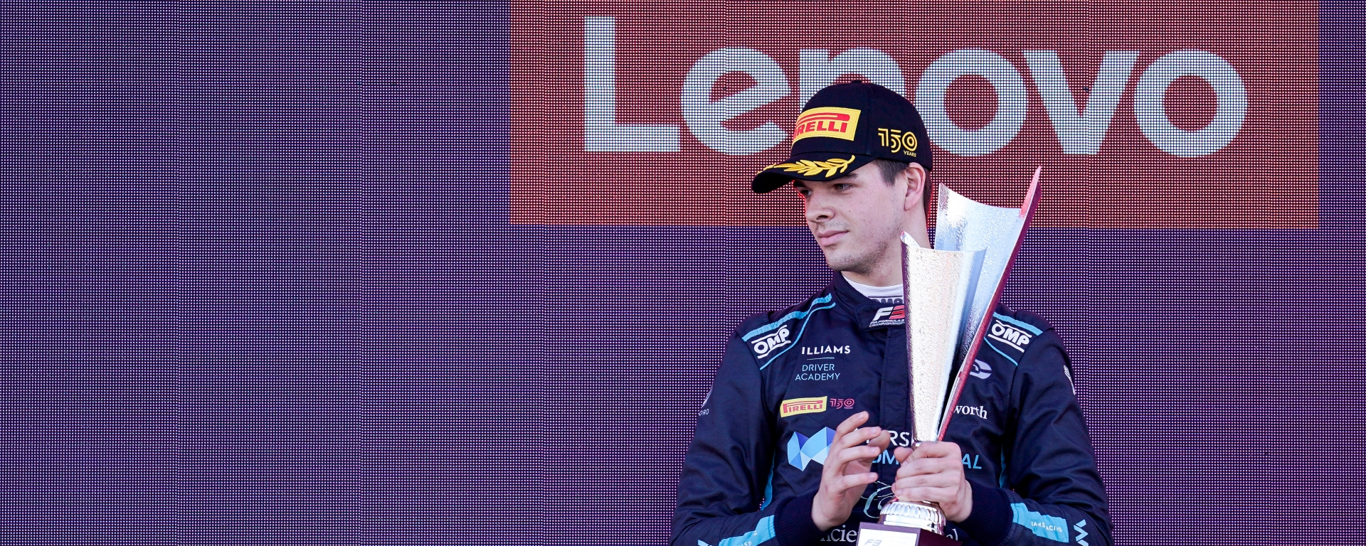 A young man (Zak O'Sullivan) in a black Pirelli 150th anniversary cap and a navy and teal racing suit holds the silver-hued second-place trophy on the podium. There is a purple screen behind him with the Lenovo logo in red.