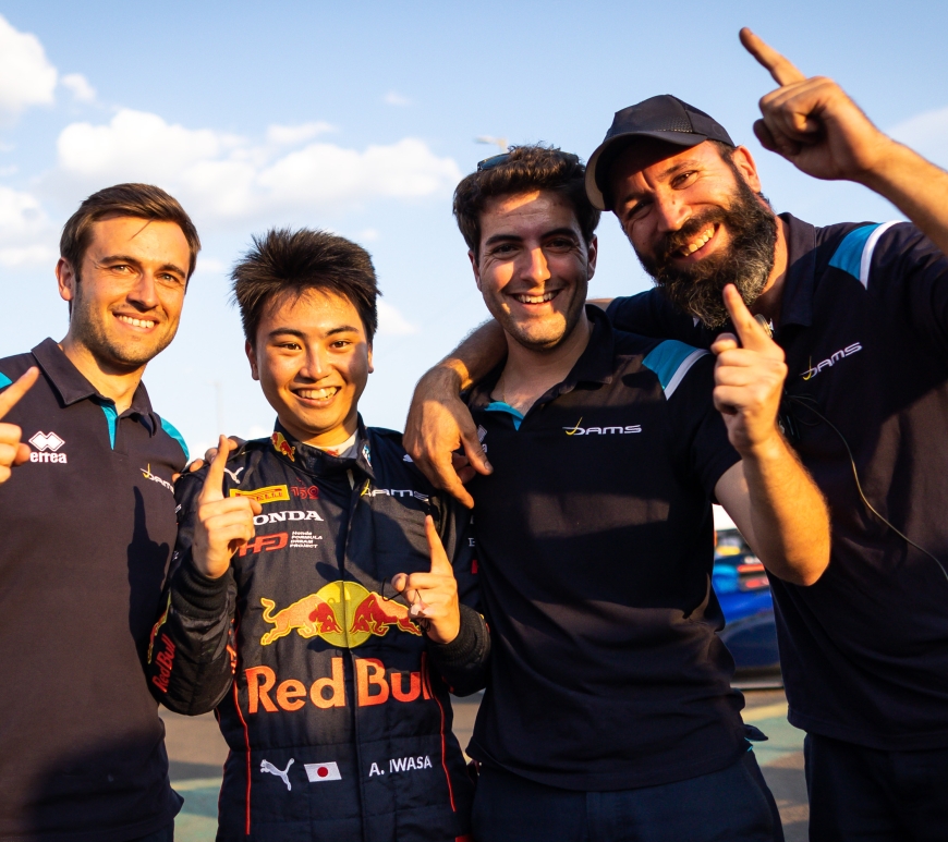 Four men in navy blue DAMS apparel smile and point their index fingers upward while draping their other arms around one another. Second from the left is DAMS driver Ayumu Iwasa, whose racing suit includes the yellow and red branding of Red Bull and his sponsors, Honda and the Honda Formula Dream Project.