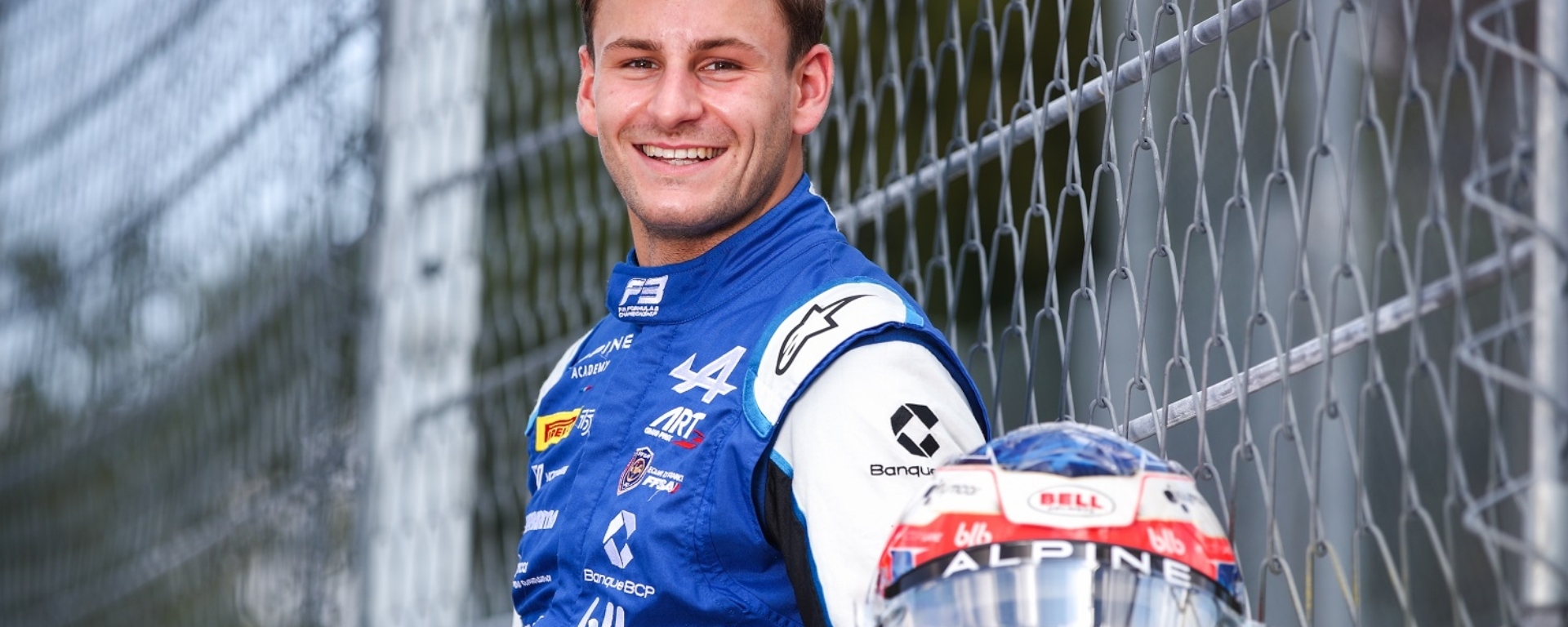 Man in blue and white racing suit standing facing to the left of the camera with head turned smiling at camera.