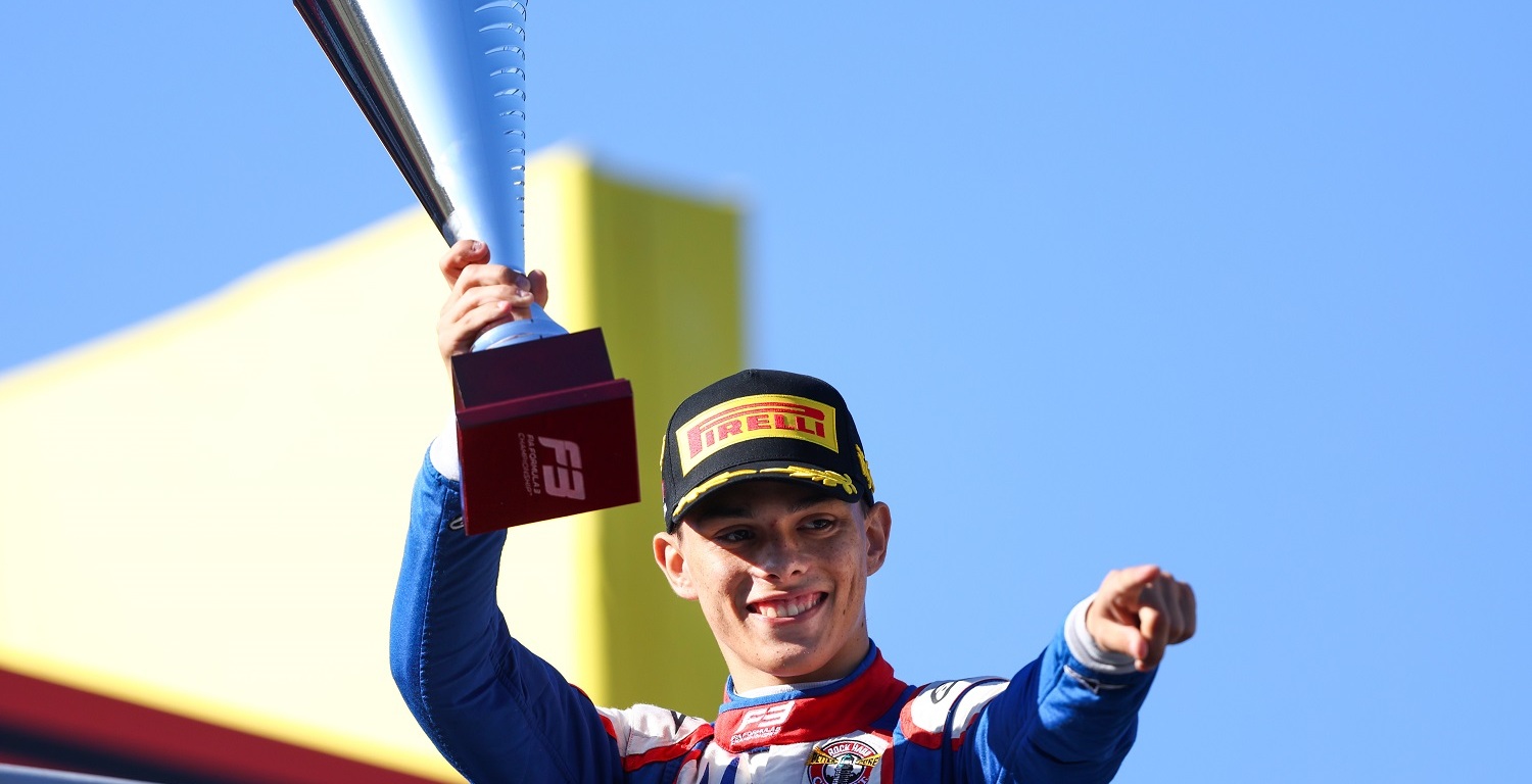 Man in red, white, and blue racing suit holding trophy above his head and pointing to the right of the camera.