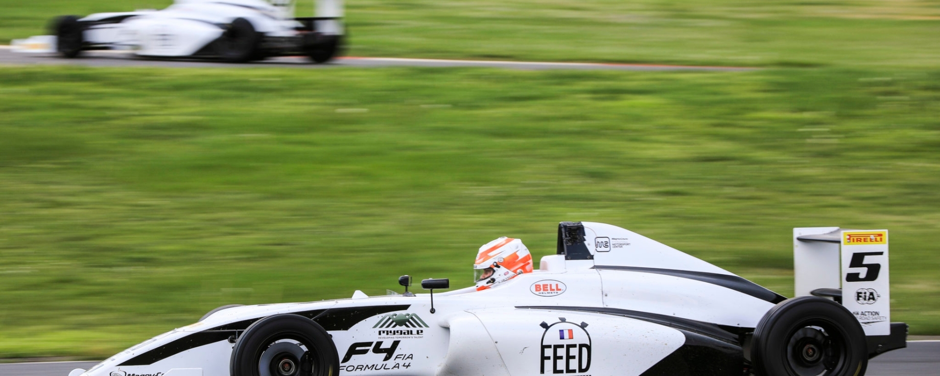 White and black formula 4 car driving to the left of the camera with another similar car behind it on a different section of the track.