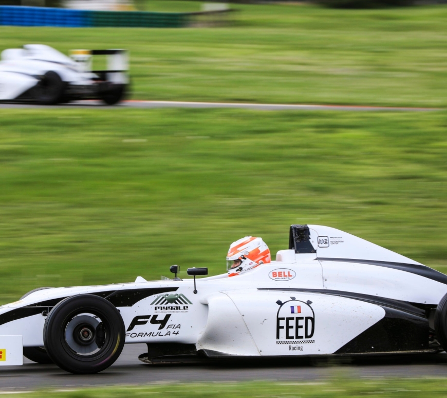 White and black formula 4 car driving to the left of the camera with another similar car behind it on a different section of the track.
