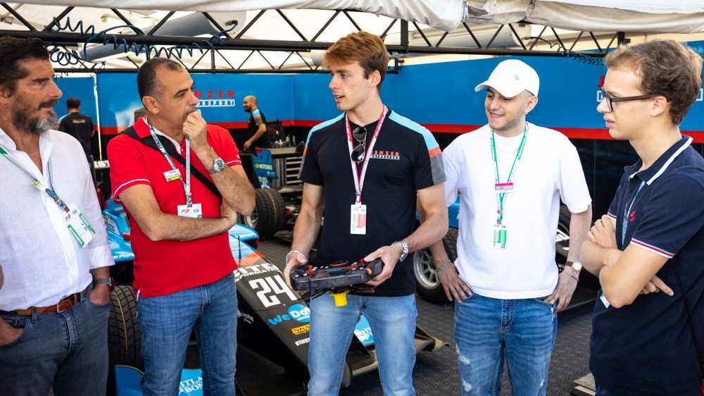 Five men in a garage with two teal, red and black racecars behind. The man in the middle, Federico Malvestiti, wears a Jenzer Motorsport–branded shirt in the same colours and holds his steering wheel as he talks with two older men to his right and two men of similar age to his left