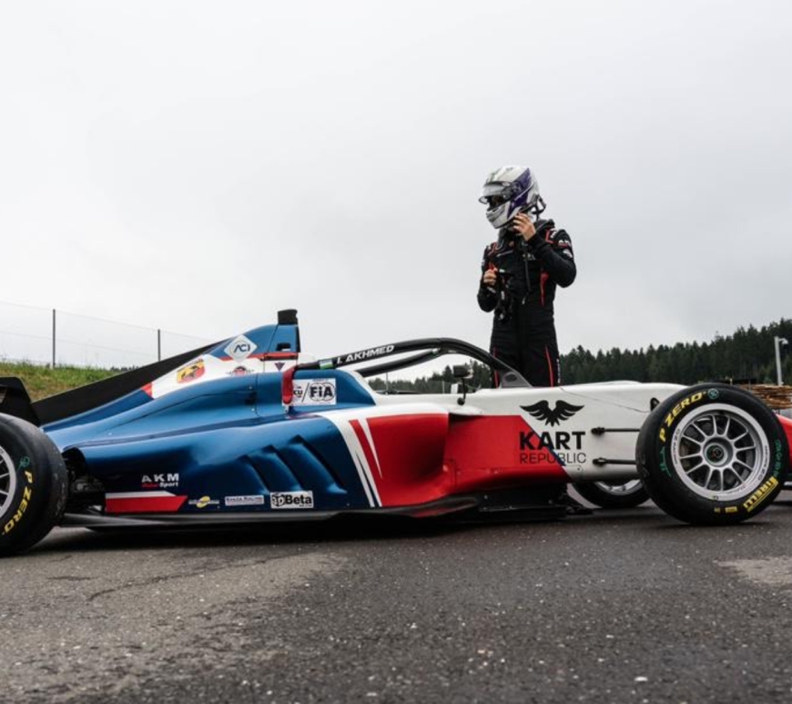 Man standing in racing suit standing behind red white and blue formula 4 car pointing sideways to the right.