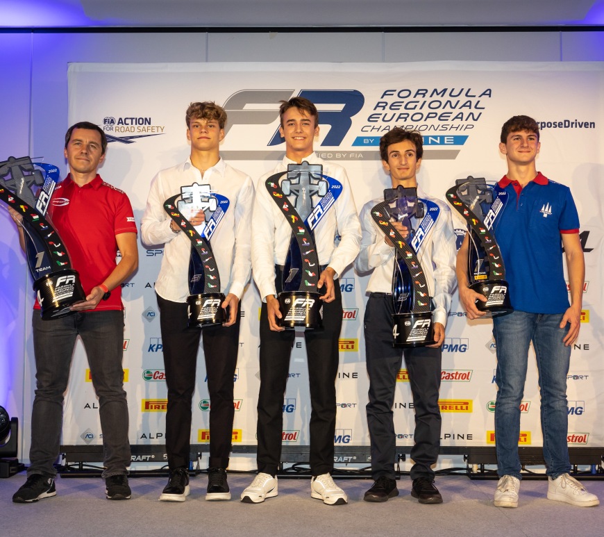 A man in a red shirt, three young men in white shirts, and a young man in a blue shirt hold trophies with a car outline at the top, the Formula Regional European Championship by Alpine logo on the base, and the outlines of all circuits on the calendar on the left edge