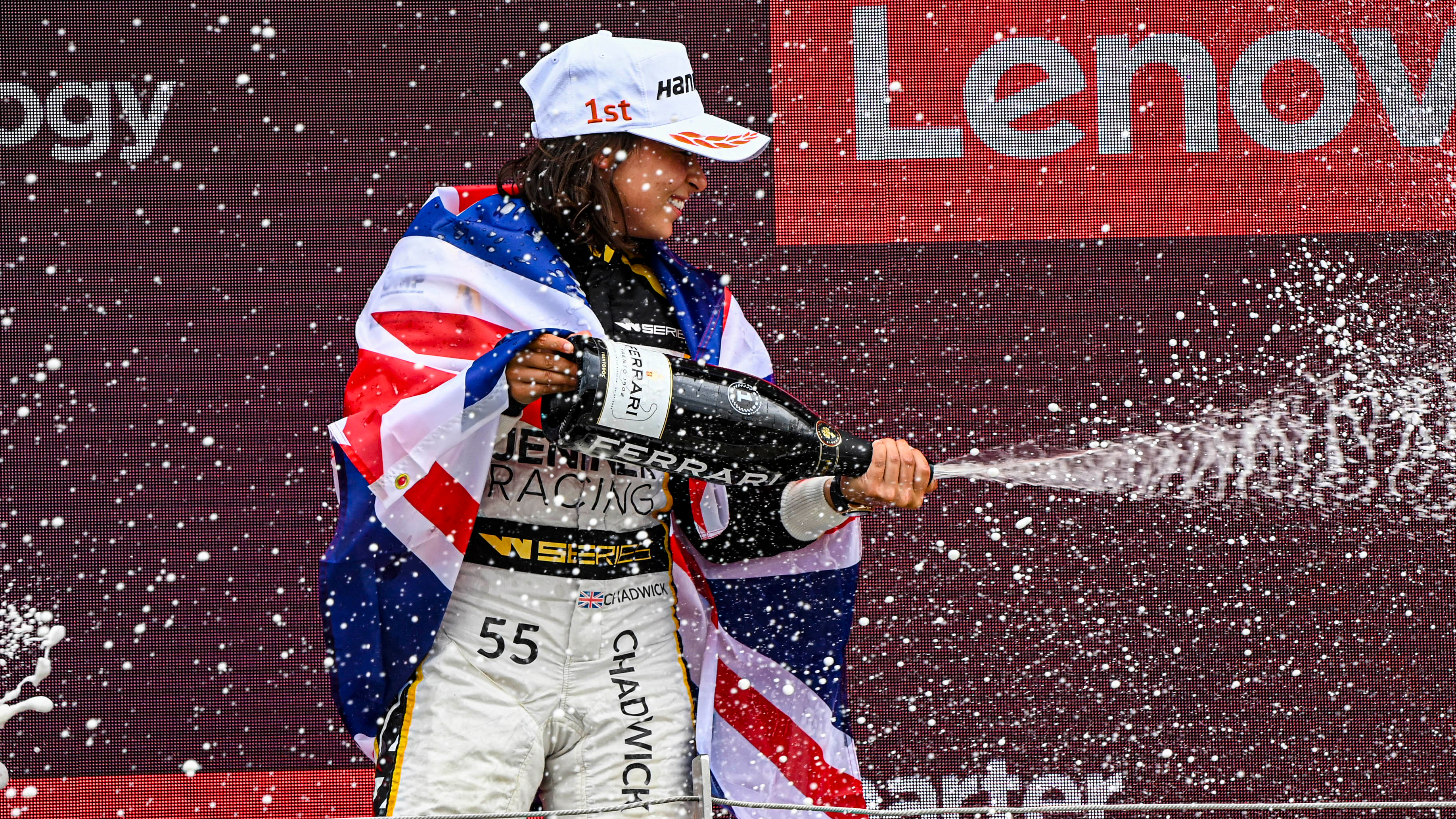 A woman with a white Hankook cap and brown hair sprays sparkling wine on the podium while draped in the Union Jack