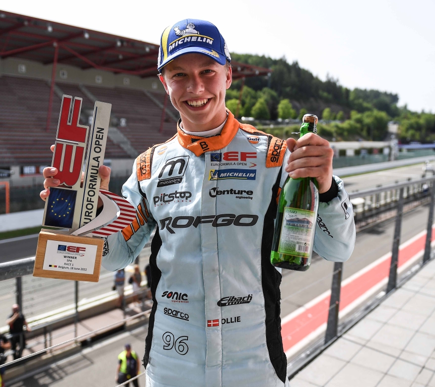 A young man in a blue cap and an orange and cyan racing suit faces the camera and grins, holding a bottle of sparkling cider in his left hand and a trophy from the Euroformula Open championship in his right hand.
