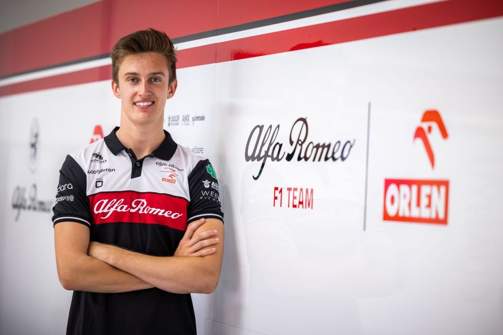 Théo Pourchaire, wearing an Alfa Romeo team shirt and crossing his arms, faces the camera and smiles. An Alfa Romeo F1 Team Orlen logo is on the wall to his right, and Alfa Romeo team sponsor logos blend into the background behind him.