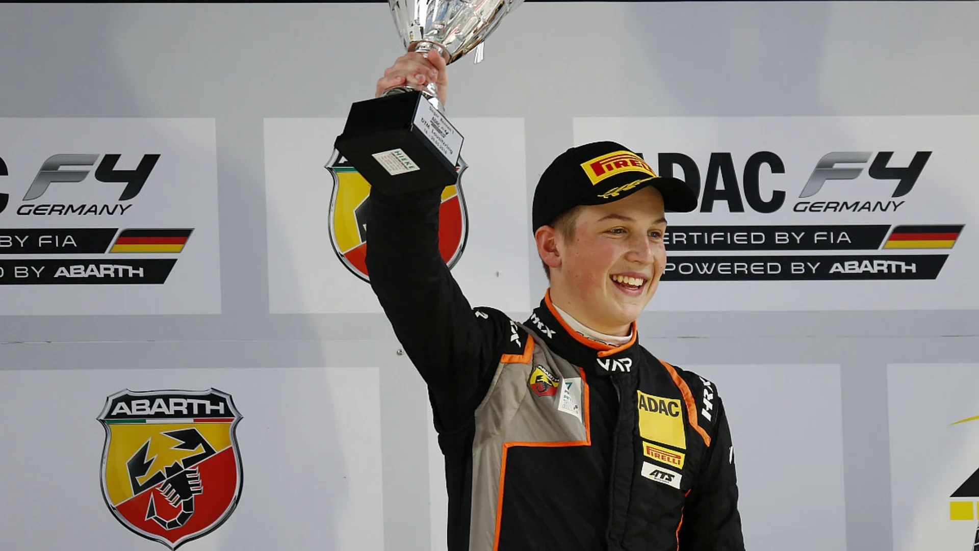 A boy in a black and orange racing suit with VAR branding smiles while looking to the lower right of the screen and lifts up a trophy that reads "Sieger Rennen 1, ADAC F4 DTM Lausitzring: 18–20.05.2018" (English: Winner, Race 1)