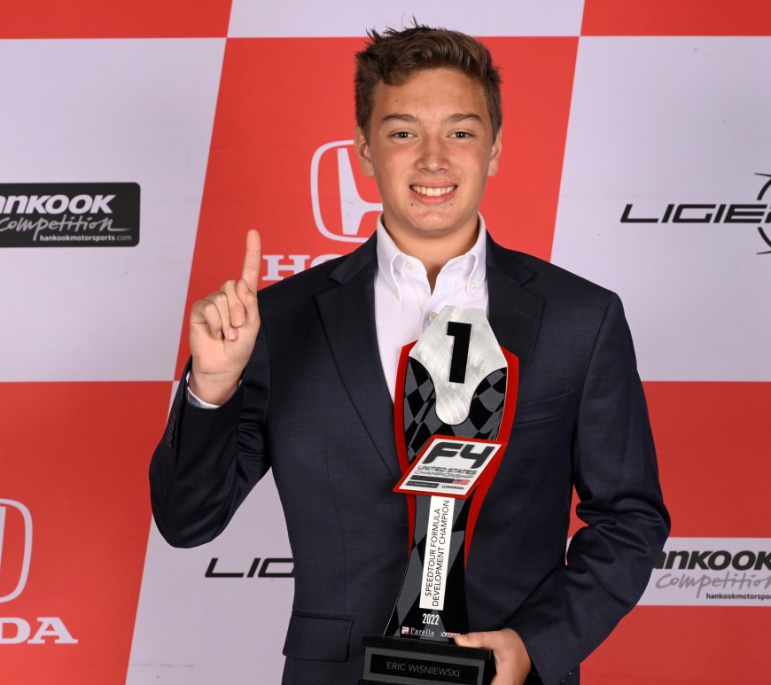 A boy with light brown hair in a suit (Eric Wisniewski) faces the camera and smiles. He lifts his right index finger in the air while holding in his left hand a trophy with the words "F4 United States Championship: SpeedTour Formula Development champion 2022" on it. Ligier, Hankook Competition, and Honda logos appear on the red and white chequered backdrop behind him.