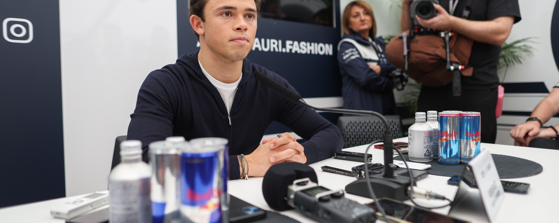 A man in a blue quarter-zip and white t-shirt sits in front of a collection of microphones and Red Bull cans.