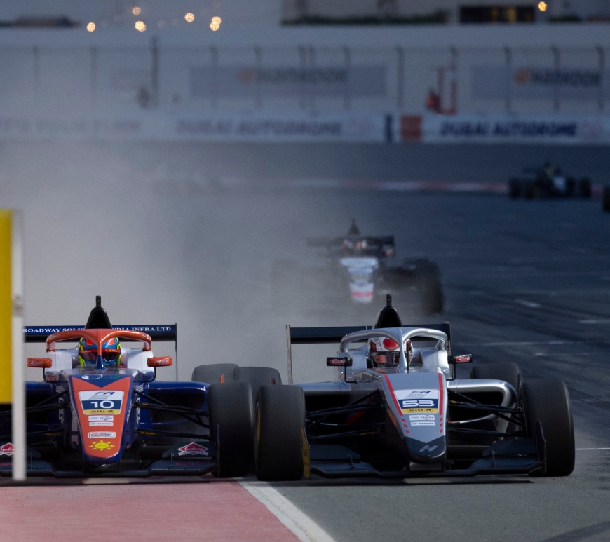 An orange, blue and white car driven by Dino Beganovic on the red tarmac alongside the pit wall, where he has been squeezed by Gabriele Minì in the silver Hitech car directly alongside him. The alteraction kicks up dust behind them