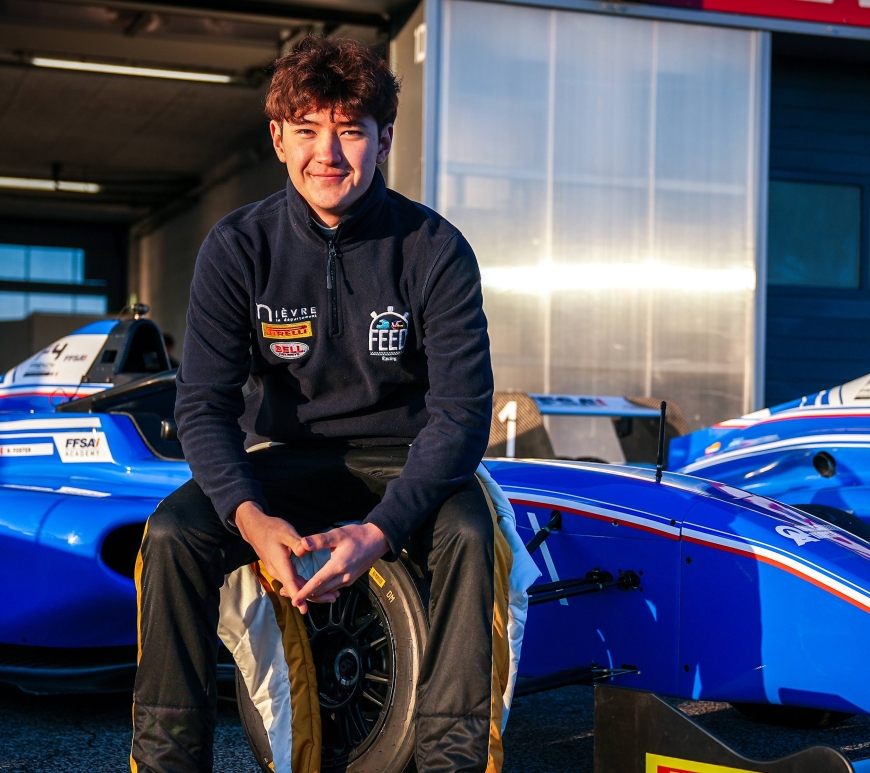 Kevin Foster, wearing a FEED Racing jacket, sits on the right-front tyre of a blue single-seater and smiles at the camera.