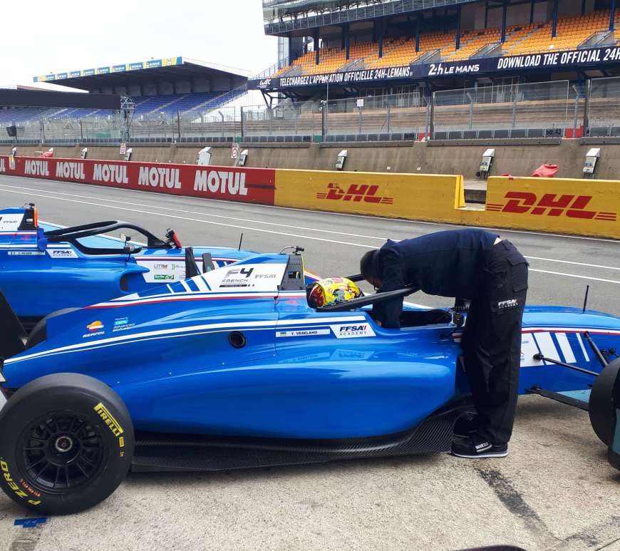 A blue and white car in pit lane, with a driver in the cockpit and an engineer leaning over to tighten belts